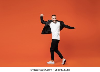 Full length side view of laughing young bearded man in basic white t-shirt black leather jacket dancing turning around to camera spreading hands isolated on orange colour background studio portrait