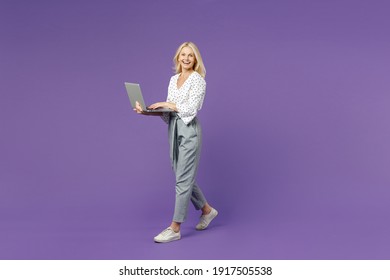 Full length side view of laughing cheerful elderly gray-haired blonde woman lady 40s 50s in white dotted blouse standing working on laptop pc computer isolated on violet background studio portrait