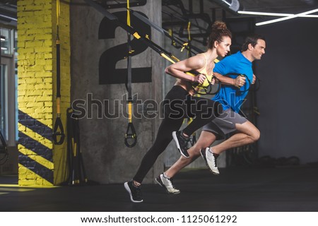 Full length side view outgoing male and concentrated girl doing exercise with functional loops. They situating in gym