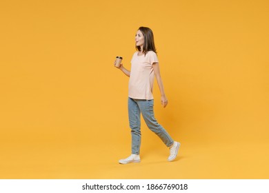 Full Length Side View Portrait Of Cheerful Funny Young Woman Wearing Pastel Pink Casual T-shirt Hold Paper Cup Of Coffee Or Tea Walking Going Looking Aside Isolated On Yellow Color Background Studio