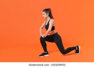Full length side view portrait of beautiful young fitness sporty woman in black sportswear posing training working out doing exercise lunge looking aside isolated on orange color background studio
