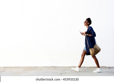 Full length side view portrait of trendy young black woman walking outdoors and listening to music on her mobile phone