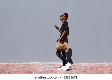 Full Length Side View Portrait Of Smiling Young Black Woman Walking Outdoors With Mobile Phone