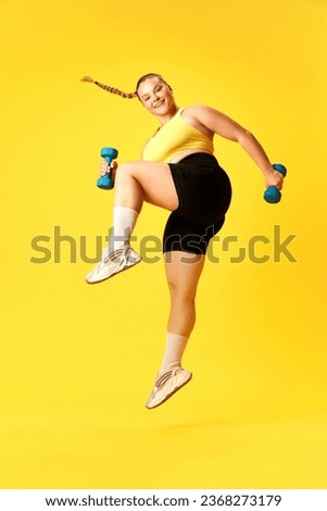Full length side view portrait of plus size woman dressed sporty training, jumping with dumbbells against over yellow background. Concept of sport, hobby, health, lifestyle, healthy eating, workout.