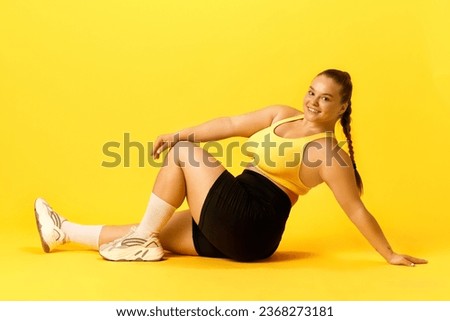 Full length side view portrait of plus size woman dressed sporty sitting on floor looking at camera against over yellow background. Concept of sport, hobby, health, lifestyle, healthy eating, workout.