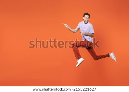 Full length side view of shocked screaming young bearded man 20s wearing casual violet t-shirt jumping pointing index fingers aside isolated on bright orange color wall background studio portrait