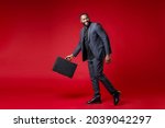 Full length side view of smiling funny young african american business man 20s wearing classic jacket suit hold in hand briefcase looking aside isolated on bright red color background studio portrait