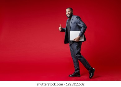 Full Length Side View Of Smiling Funny Young African American Business Man 20s In Classic Jacket Suit Hold Laptop Pc Computer Showing Thumb Up Isolated On Bright Red Color Background Studio Portrait