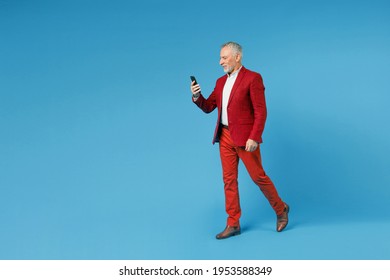 Full Length Side View Of Smiling Elderly Gray-haired Mustache Bearded Business Man Wearing Red Jacket Suit Using Mobile Cell Phone Typing Sms Message Isolated On Blue Color Background Studio Portrait