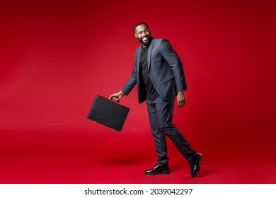 Full Length Side View Of Smiling Funny Young African American Business Man 20s Wearing Classic Jacket Suit Hold In Hand Briefcase Looking Aside Isolated On Bright Red Color Background Studio Portrait