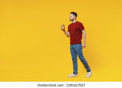 Full length side view smiling young man 20s in red t-shirt casual clothes hold takeaway delivery craft paper brown cup coffee to go walk isolated on plain yellow color wall background studio portrait.