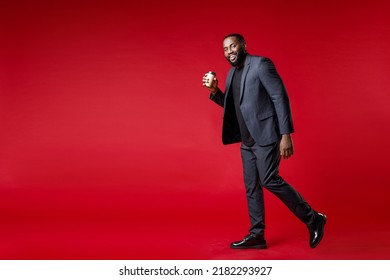 Full Length Side View Of Smiling Young African American Business Man 20s In Classic Jacket Suit Hold Paper Cup Of Coffee Or Tea Look Camera Goes Isolated On Bright Red Color Background Studio Portrait