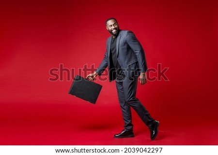 Full length side view of smiling funny young african american business man 20s wearing classic jacket suit hold in hand briefcase looking aside isolated on bright red color background studio portrait
