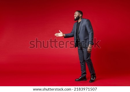 Full length side view of smiling young african american business man 20s wearing classic jacket suit standing with outstretched hand for greeting isolated on red color background studio portrait