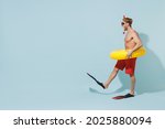 Full length side view young man in red shorts swimsuit diving mask near pool hold inflatable ring flippers raise up leg isolated on pastel blue background. Summer vacation sea rest sun tan concept.