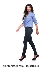 Full Length Side View Of A Young Casual Woman Walking Forward While Looking Into The Camera. On White Background