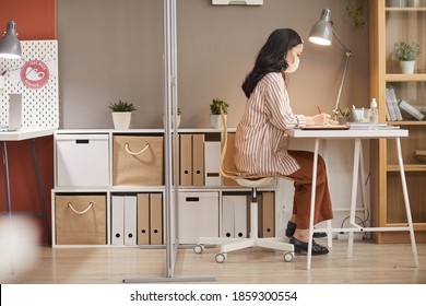 Full Length Side View At Young Asian Woman Wearing Mask In Office While Working At Desks In Cubicle Post Pandemic, Copy Space