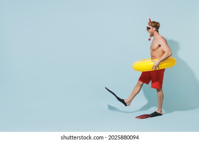 Full length side view young man in red shorts swimsuit diving mask near pool hold inflatable ring flippers raise up leg isolated on pastel blue background. Summer vacation sea rest sun tan concept.