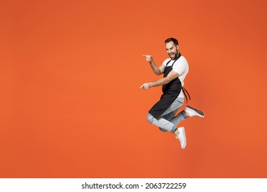 Full Length Side View Young Man Barista Bartender Employee In Black Apron White T-shirt Work In Coffee Shop Jump High Point Finger Aside On Area Isolated On Orange Background. Small Business Startup