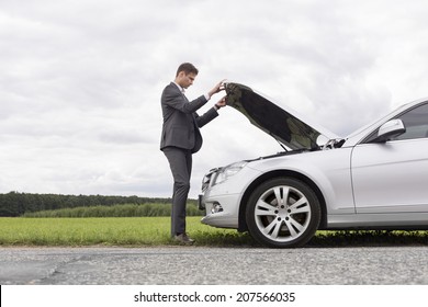 Full length side view of young businessman opening broken down car hood at countryside