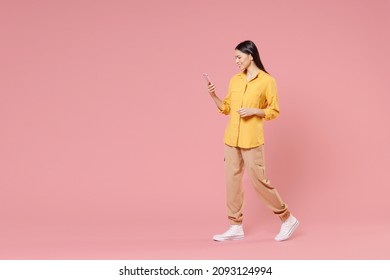 Full length side view of young smiling brunette positive nice attractive latin woman 20s wearing yellow shirt hold mobile cell phone walking isolated on pastel pink color background studio portrait