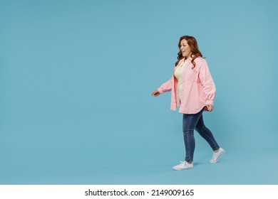 Full Length Side View Young Fun Smiling Redhead Chubby Overweight Woman 30s Wearing In Pink Shirt Jeans Casual Clothes Walking Going Looking Aside Isolated On Pastel Blue Background Studio Portrait.