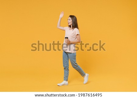 Full length side view young woman in pink casual t-shirt hold takeaway delivery craft paper brown cup coffee to go waving greet with hand as notices someone isolated on yellow color background studio
