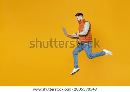 Full length side view young strong powerful fun caucasian man 20s years old in orange vest mint sweatshirt jump high hold laptop pc computer chat online isolated on yellow background studio portrait