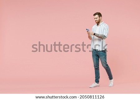 Full length side view young smiling unshaven man 20s in blue striped shirt hold mobile cell phone chat online browsing internet walk isolated on pastel pink background studio. Tattoo translate fun.