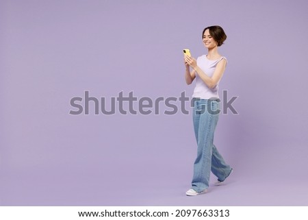 Full length side view young smiling happy fun brunette woman 20s in white t-shirt using mobile cell phone chat online browsing internet walk go isolated on pastel purple background studio portrait.