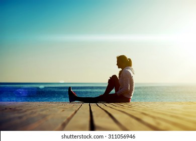 Volllange Silhouette junger Läufer, die auf dem Holzsteg auf dem Ozean sitzen und sich nach einem Lauf am schönen Abend ausruhen, Sportlerin mit guter Figur, die sich nach einem aktiven Rundgang im Freien erholt