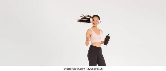 Full length of smiling attractive asian female athelte, girl doing fitness workout in gym, sportswoman or runner drinking water from bottle, listening music in wireless headphones, tossing hair - Shutterstock ID 2208595105