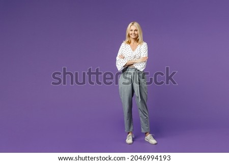 Full length of smiling attractive beautiful elderly gray-haired blonde woman lady 40s 50s in white dotted blouse holding hands crossed looking camera isolated on violet background studio portrait