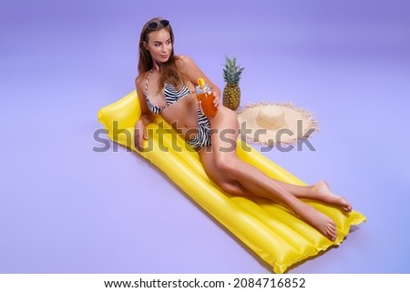 Full length of smiling beautiful caucasian woman who wears in bikini lyes on inflatable yellow matress while holds cocktail on purple background . Summer concept. Travel concept. Relax concept 