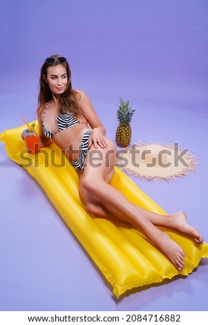 Full length of smiling beautiful caucasian woman who wears in bikini lyes on inflatable yellow matress while holds cocktail on purple background . Summer concept. Travel concept. Relax concept 