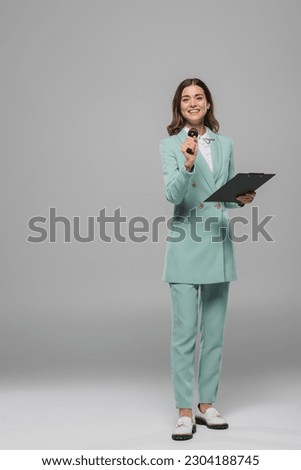 Full length of smiling and brunette event host in blue suit holding microphone and clipboard while looking at camera and standing on grey background