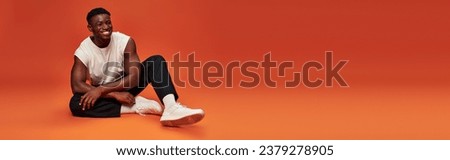 full length of smiling fashionable african american man sitting on red and orange backdrop, banner