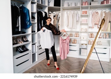 Full Length Of Smiling Female In Trendy Clothes Standing In White Walk In Closet With Many Shelves Near Mirror And Selecting Outfit At Home