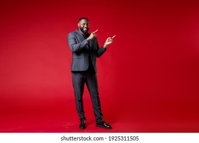 Full Length Of Smiling Handsome Young African American Business Man 20s Wearing Classic Jacket Suit Standing Pointing Index Fingers Aside Isolated On Bright Red Color Wall Background Studio Portrait