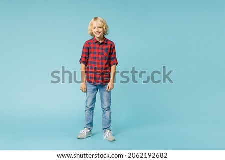 Full length of smiling little curly kid boy 10s years old in basic red checkered shirt standing looking camera isolated on blue color background children studio portrait. Childhood lifestyle concept