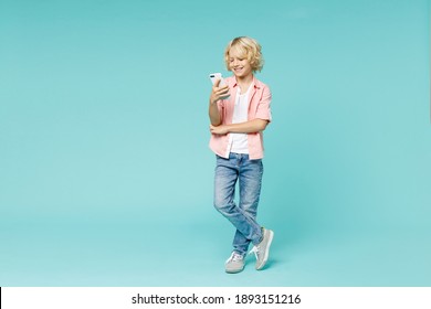 Full Length Of Smiling Little Kid Boy 10s In Pink Shirt Using Mobile Cell Phone Typing Sms Message Isolated On Blue Turquoise Color Background Children Studio Portrait. Childhood Lifestyle Concept