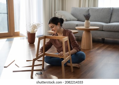 Full length smiling millennial generation indian woman sitting on warm heated floor, constructing wooden furniture in modern living room, fixing broken chair or table, improving interior at home. - Shutterstock ID 2082214333