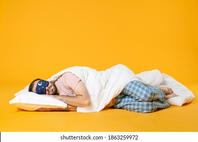 Full length smiling pretty young bearded man in pajamas home wear sleep mask lying with pillow blanket isolated on bright yellow colour background studio portrait. Relax good mood lifestyle concept