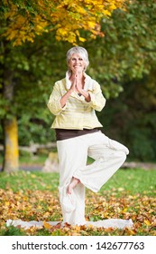 Full Length Of Smiling Senior Woman With Arms Outstretched Standing On One Leg While Doing Yoga In Park