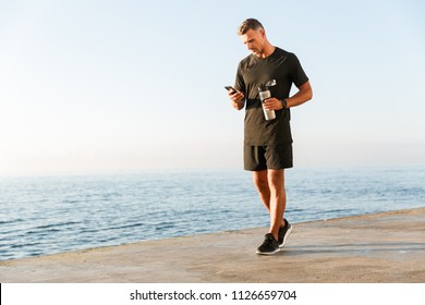 Full length of smiling sportsman using mobile phone while standing at the beach and holding bottle of water - Powered by Shutterstock