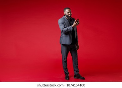 Full length of smiling successful young african american business man 20s in classic jacket suit using mobile cell phone typing sms message isolated on bright red color background studio portrait