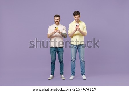 Full length smiling two young happy men friends together stand in casual t-shirt using mobile cell phone chat online isolated on purple background studio People lifestyle concept. Tattoo translate fun