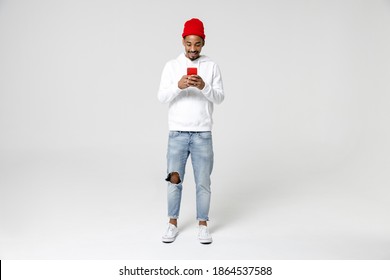 Full Length Of Smiling Young African American Man 20s Years Old In Casual Streetwear Hoodie Standing Using Mobile Cell Phone Typing Sms Message Isolated On White Colour Background, Studio Portrait