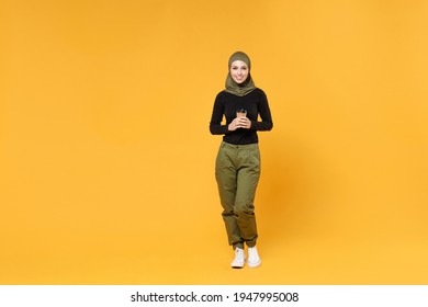 Full length of smiling young arabian muslim woman in hijab black green clothes hold paper cup of coffee or tea isolated on yellow color background studio portrait. People religious lifestyle concept
