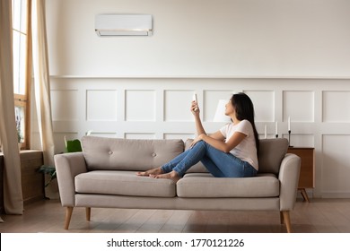 Full length smiling young asian ethnic woman sitting on cozy sofa, turning on off air conditioner with remote controller, setting indoors temperature preference, managing cooling system at home. - Shutterstock ID 1770121226